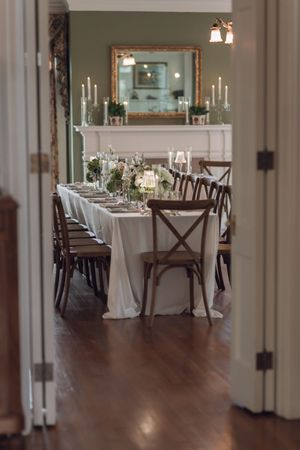 Elegant wedding reception room with long white-draped table, wooden cross-back chairs, floral centerpieces, and candles by a fireplace.