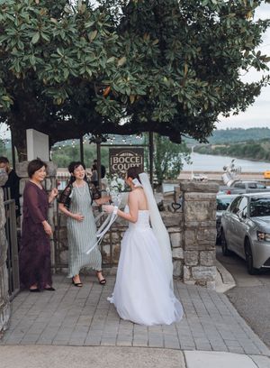 Bride in white gown with veil greets guests near Bocce Court sign with waterfront view.