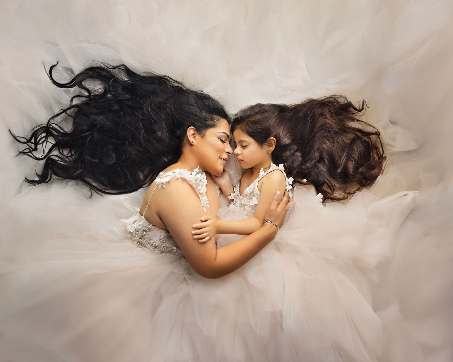 Mother and daughter in matching white tulle dresses lying together, sharing a tender embrace with flowing dark hair spread out.
