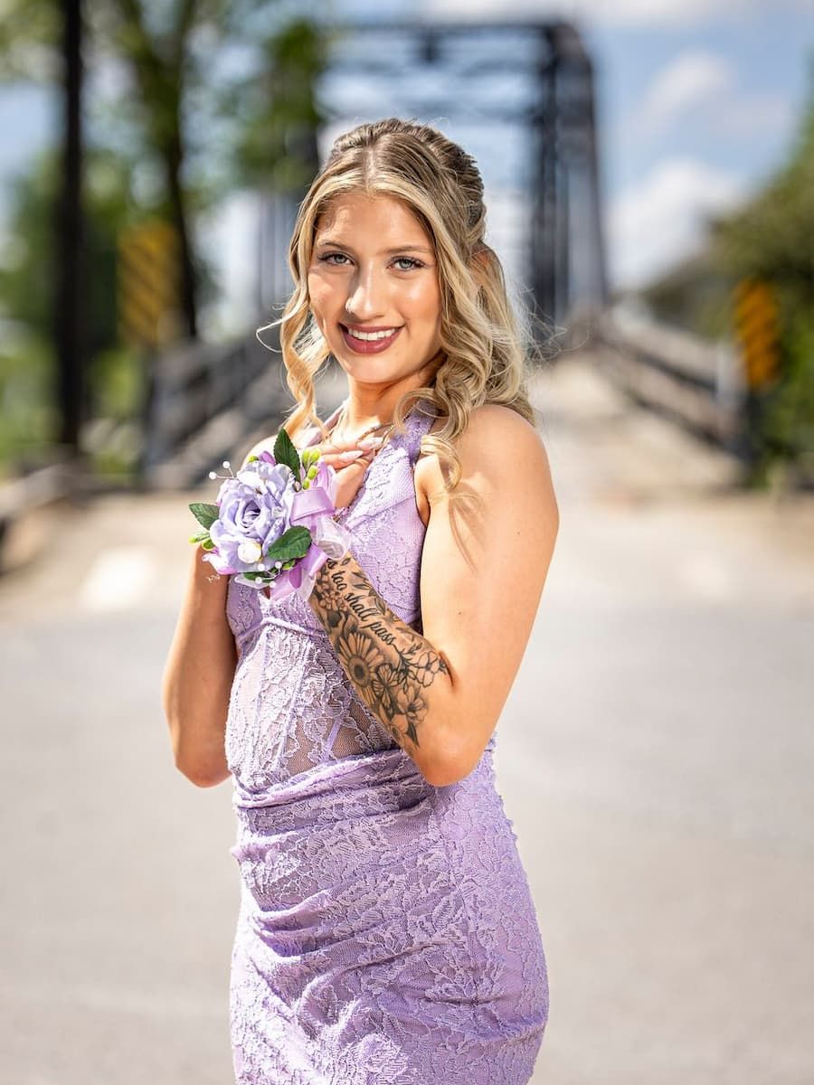 A couple in prom attire poses by a river under a bridge, she in lavender gown, he in dark suit with purple tie.