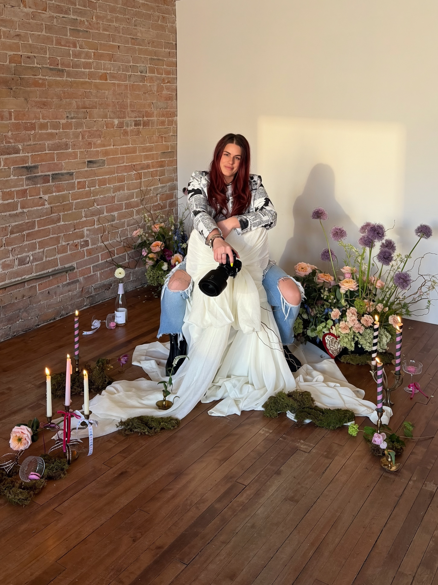 Woman with red hair sits among flowers and candles in a styled editorial shoot with brick wall backdrop.