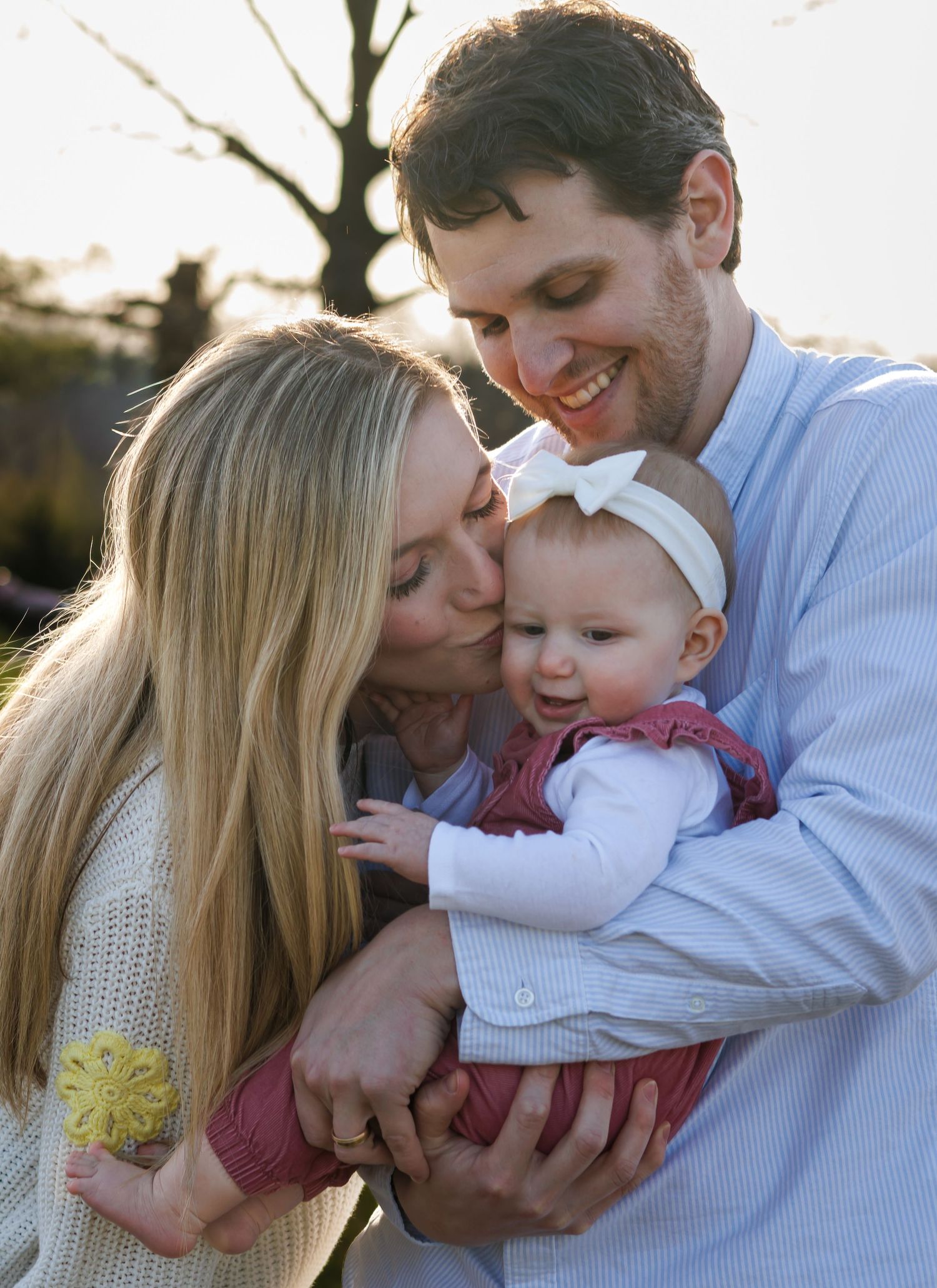 Happy family portrait with parents kissing baby girl outdoors at golden hour.