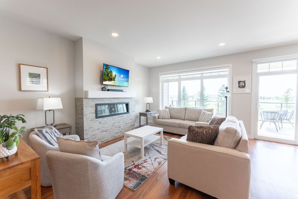 Bright modern living room with hardwood floors, stone fireplace, wall-mounted TV, neutral sofas, and large windows with outdoor views.
