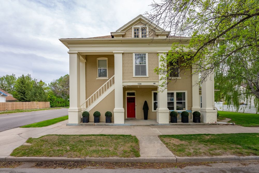Two-story beige colonial home with red front door, white columns, and manicured shrubs on a corner lot.