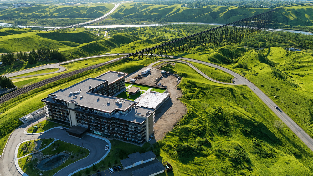 Aerial view of a modern hotel building nestled among lush green rolling hills with winding roads and a bridge in the background.