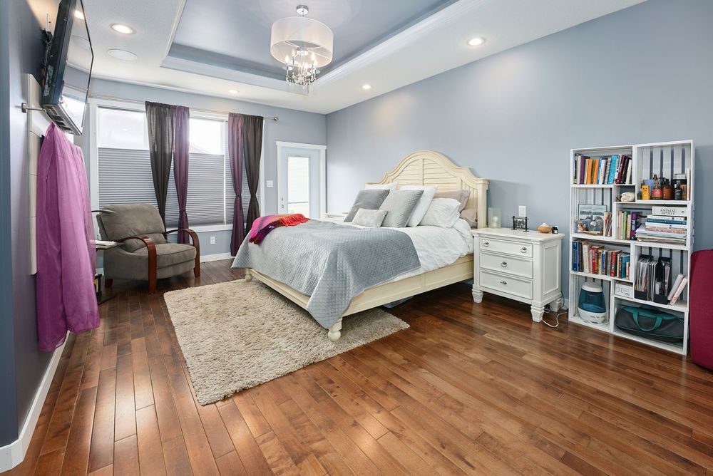 Spacious bedroom with hardwood floors, tray ceiling, white bed frame, bookshelf, and natural light from large windows.