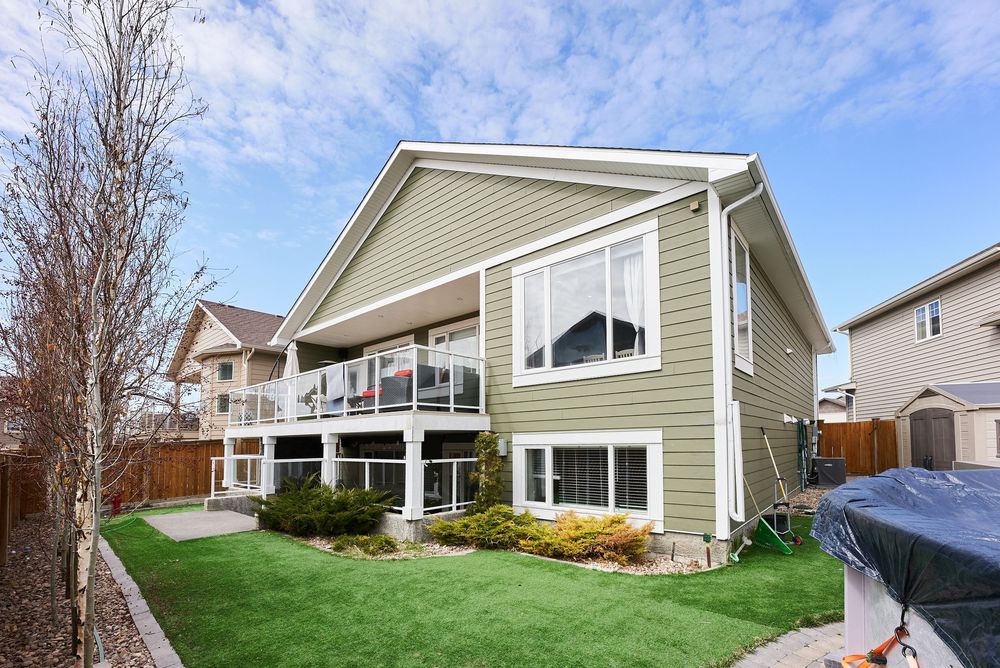 Two-story green home with large deck, white trim, manicured lawn, and landscaped yard under a partly cloudy sky.
