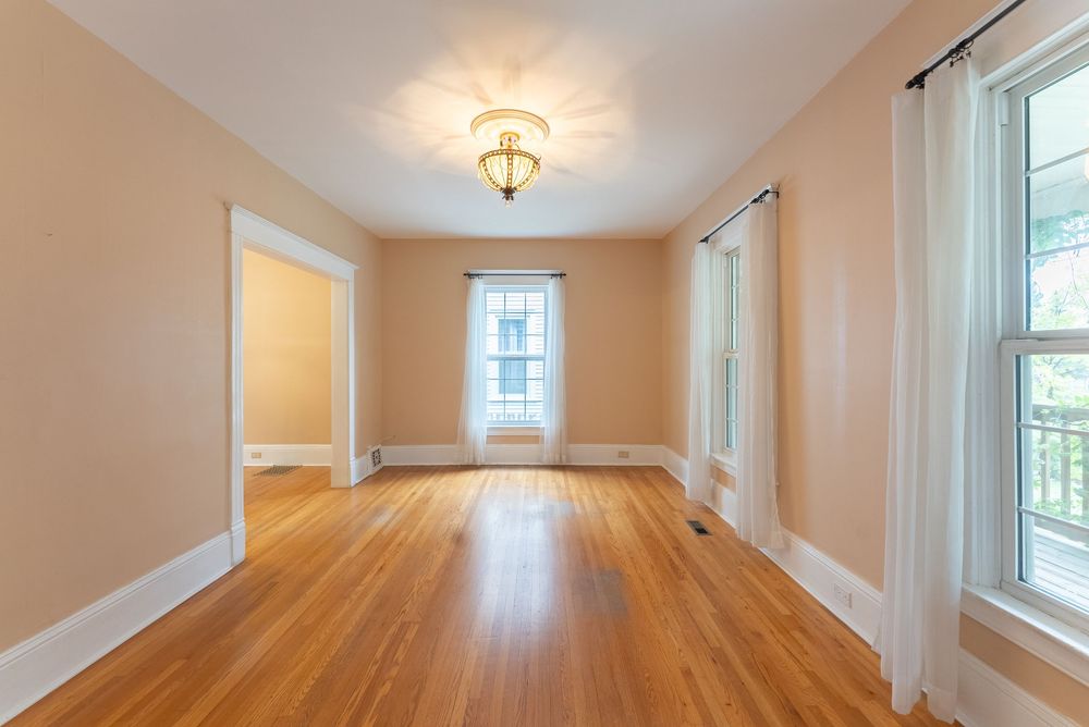 Bright empty room with hardwood floors, peach walls, white trim, a ceiling light fixture, and large windows.