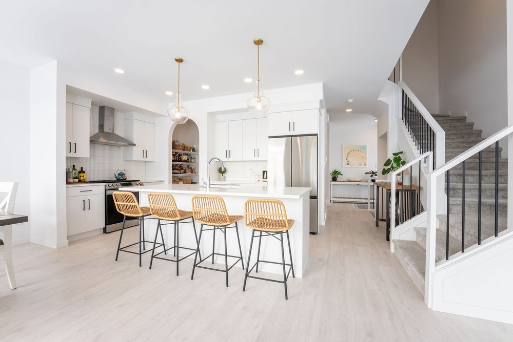 Open-concept white kitchen with large island, rattan bar stools, gold pendant lights, and adjacent staircase.