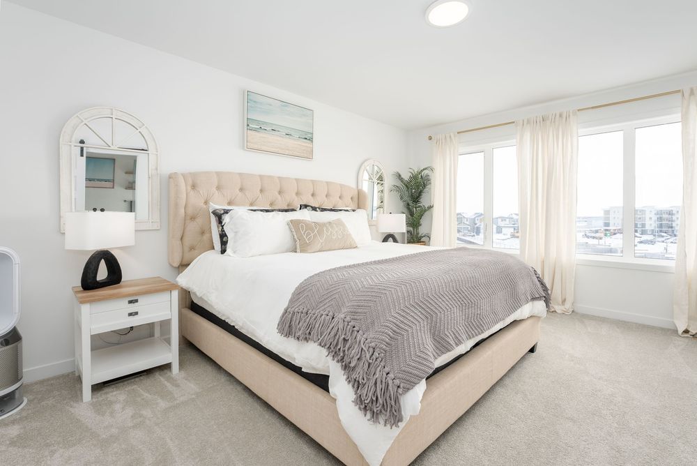 Bright master bedroom with tufted beige headboard, neutral decor, plush carpet, and large windows with natural light.