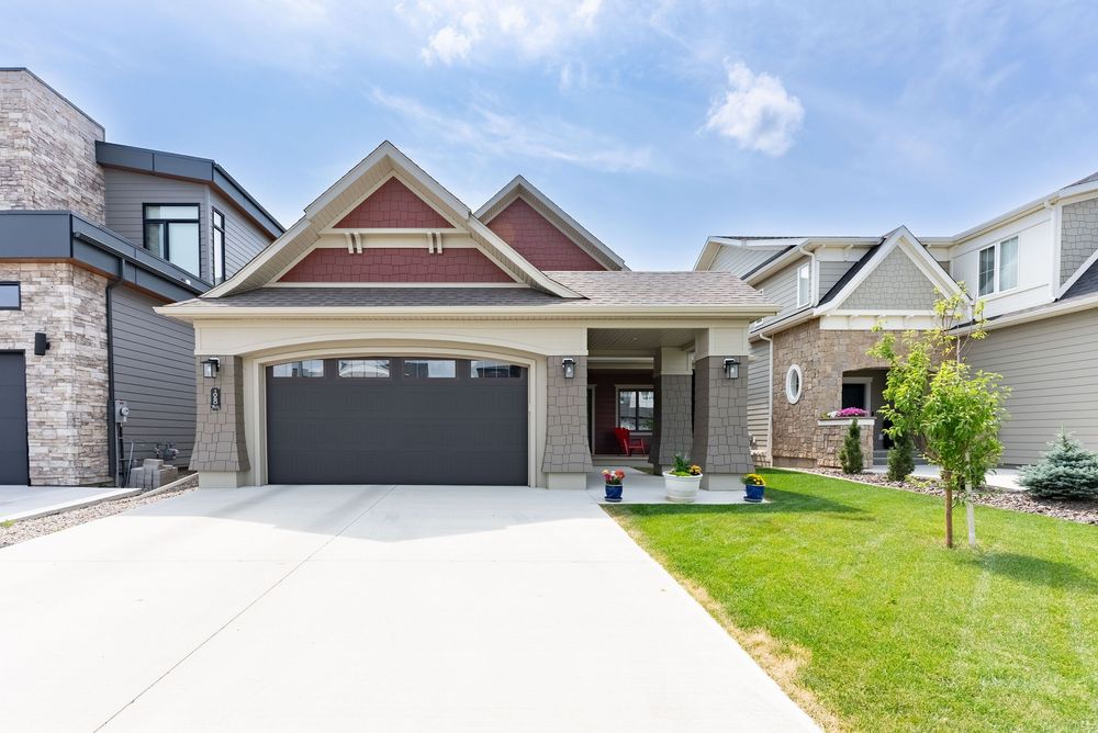 Craftsman-style home with red accent gables, dark double garage door, manicured lawn, and stone facade on a sunny day.