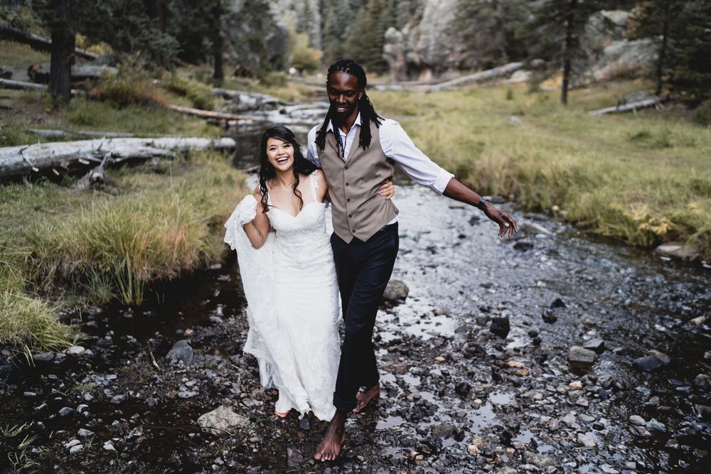 Happy interracial newlywed couple laughing while crossing a rocky stream in a lush forest setting in Jemez Wilderness