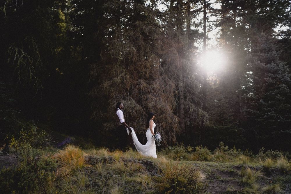 Groom lifts bride in white gown in sunlit forest clearing with golden light streaming through tall pines in Jemez Wilderness