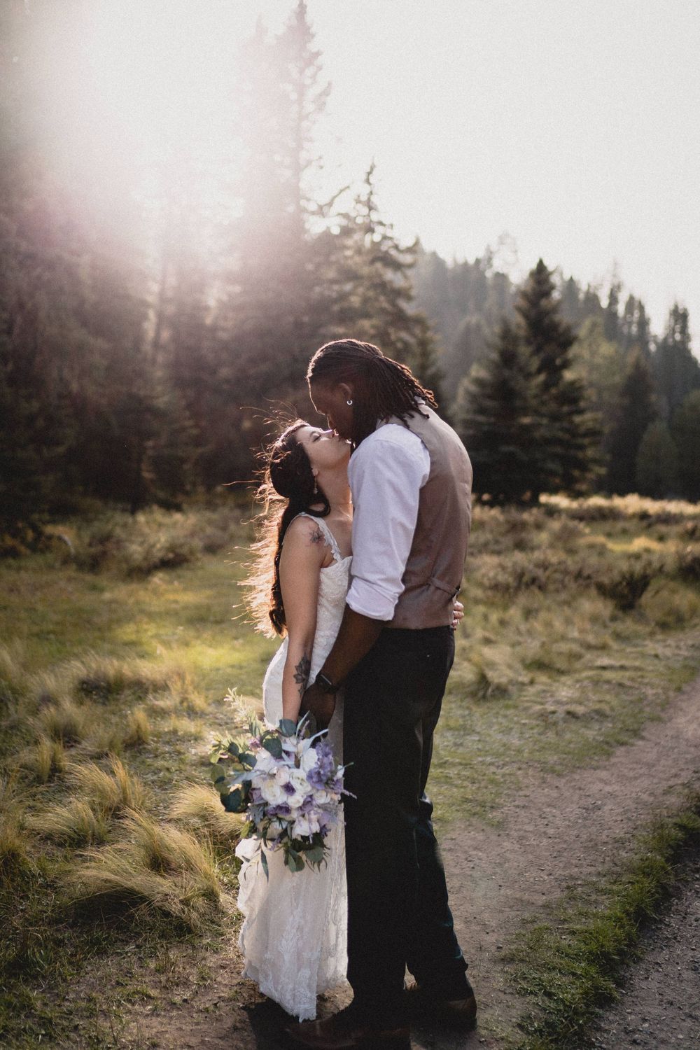 Bride and groom sharing a romantic kiss on a rustic path with golden sunlight filtering through pine trees in Jemez Wilderness