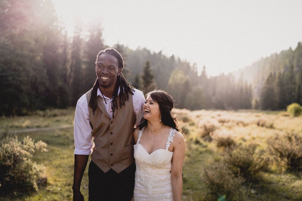 Joyful interracial newlywed couple laughing together in a sunlit meadow surrounded by evergreen forest in Jemez Wilderness