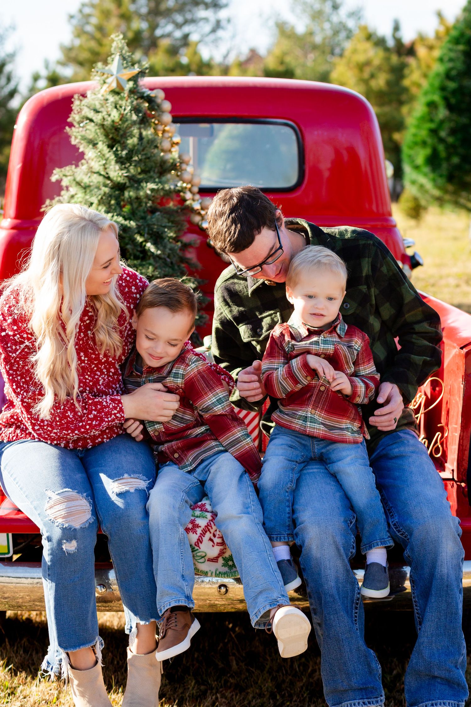 Family of four in matching plaid sits on red truck tailgate at Christmas tree farm on sunny day.