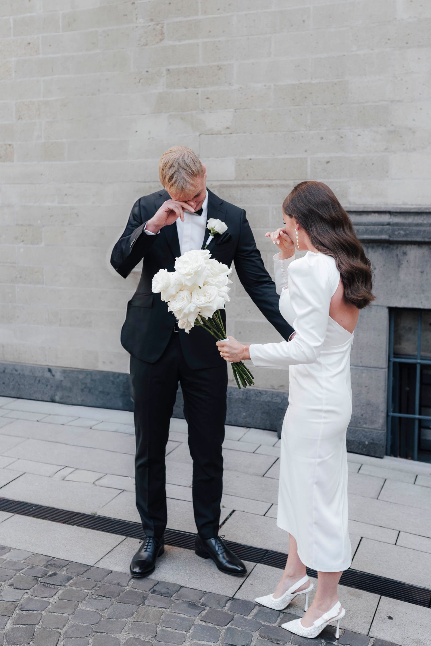 Ein gerührtes Brautpaar wischt sich die Tränen weg während des First Looks. Die Braut in einem weißen Midikleid und Brautstrauß mit Rosen.