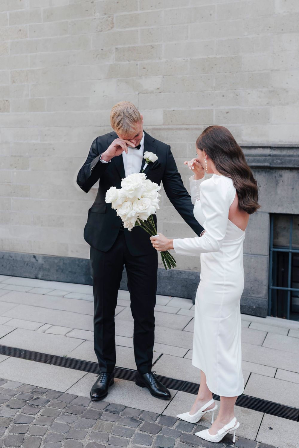 Emotional groom in black suit wiping tears as bride in white midi dress holds bouquet of white peonies outdoors.