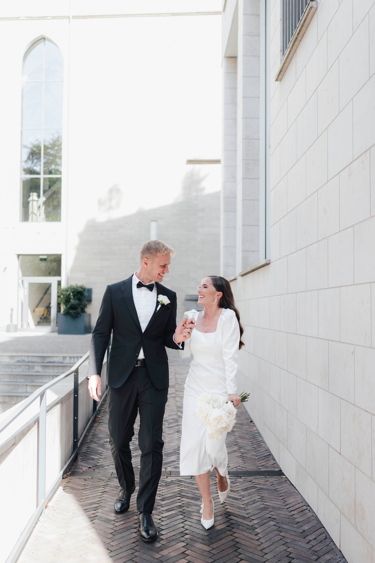 A bride and groom walk together laughing outside a white church, she holds a bouquet wearing a midi dress, he wears a black tuxedo.