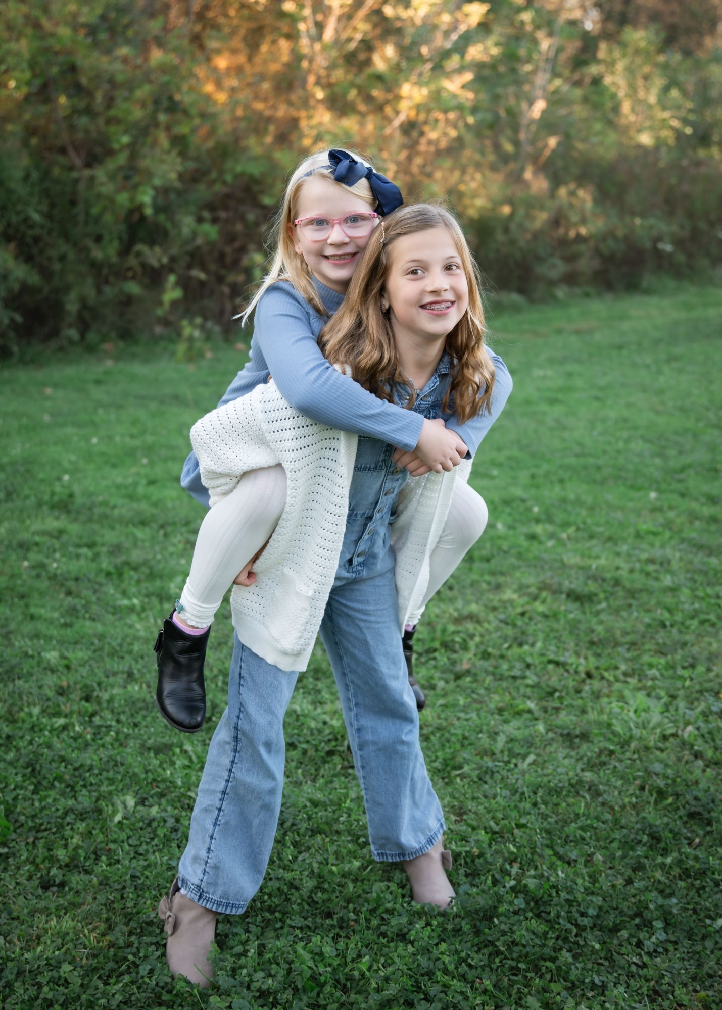 Two smiling girls share a piggyback ride outdoors on green grass, wearing casual blue outfits with trees in the background.