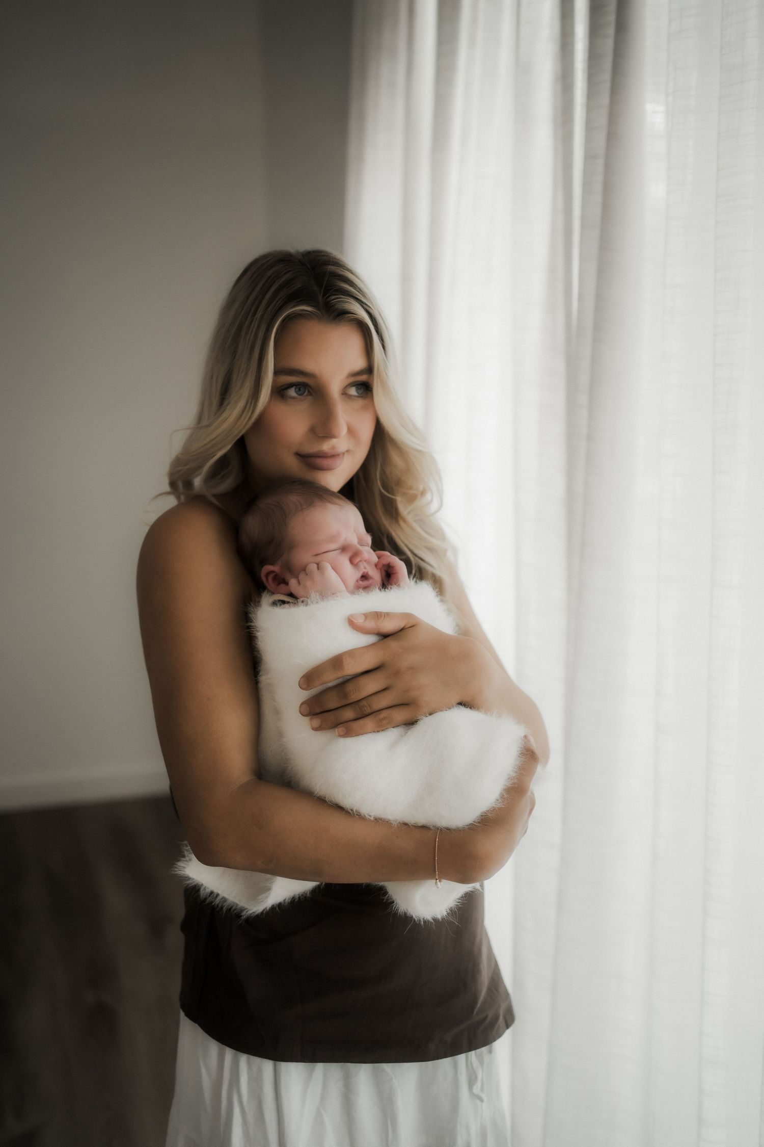 Young blonde mother tenderly holds sleeping newborn baby wrapped in white fluffy blanket near bright window in Buxton, Victoria