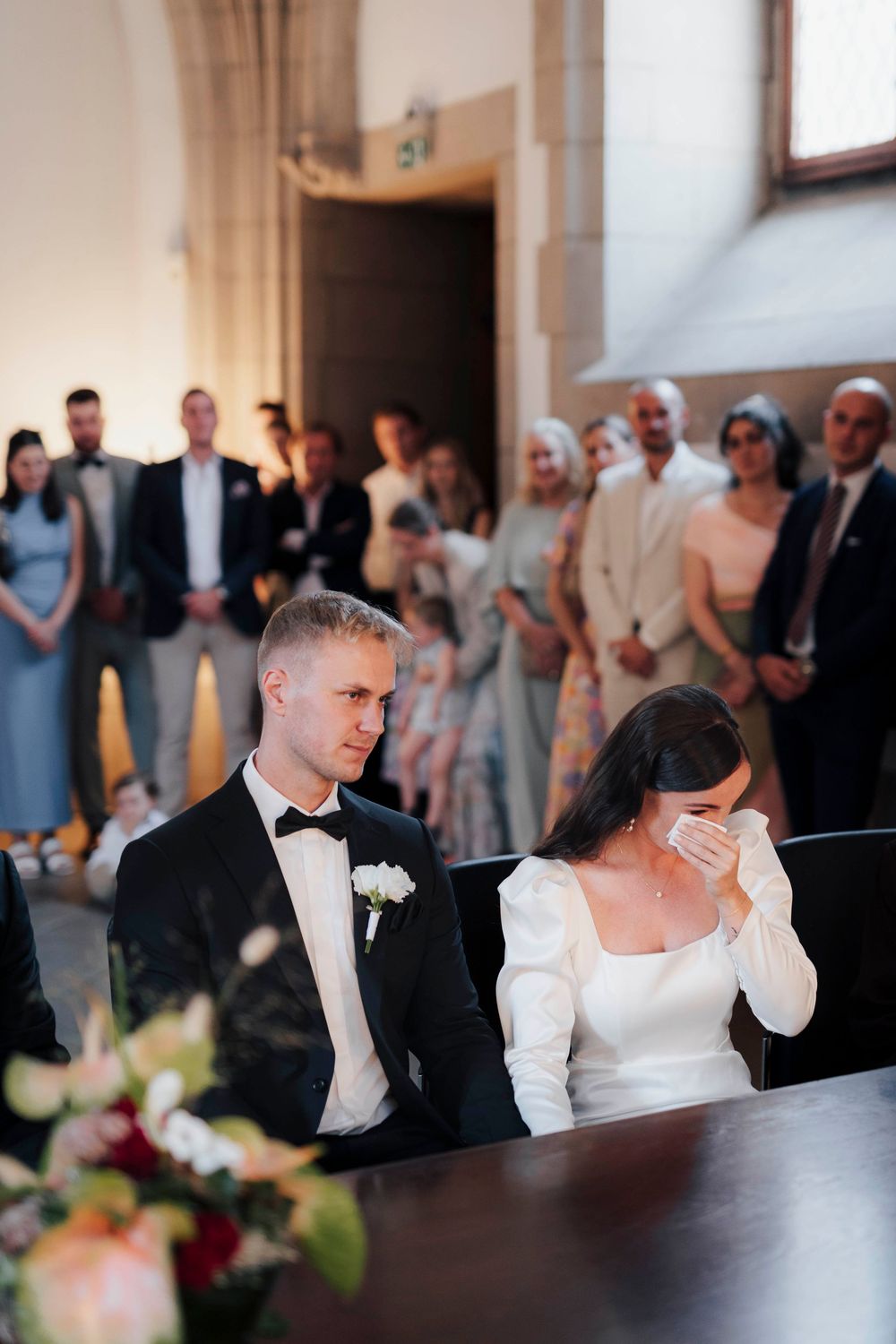 Emotional bride wiping tears at wedding ceremony while groom sits beside her, guests standing in background.