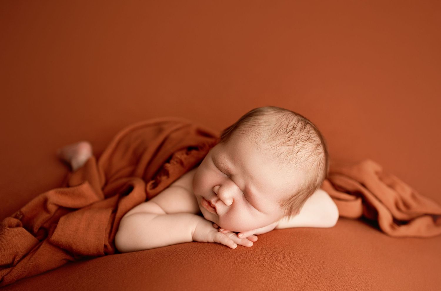 Sleeping newborn baby posed on rust-orange blanket with hands tucked under chin in professional photography studio.