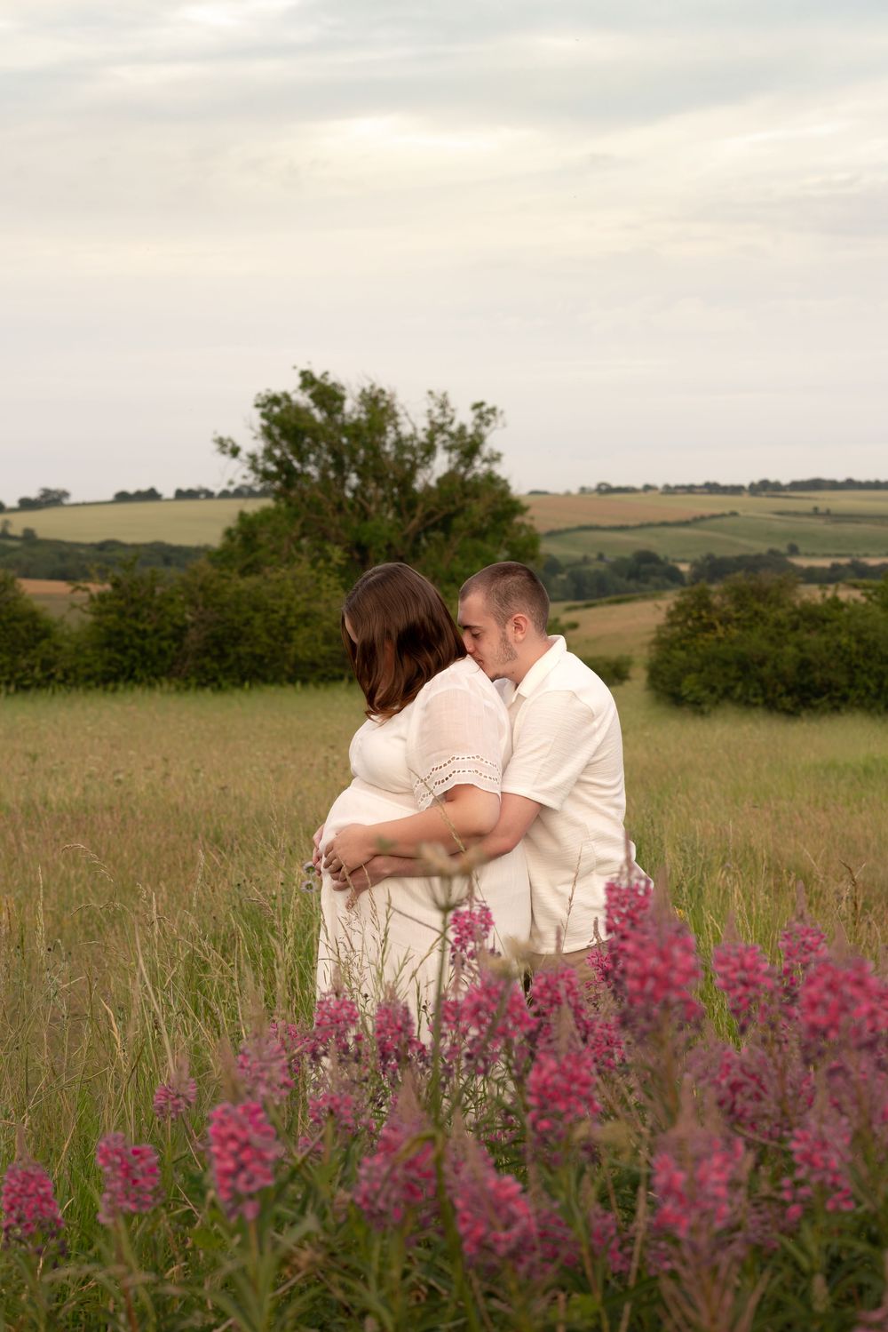 Couple embracing in a wildflower meadow during maternity photoshoot, with rolling green hills and trees in background.