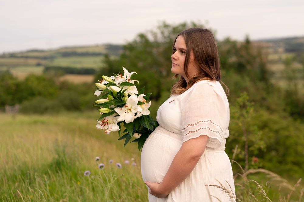 Pregnant woman in white dress holding white lilies in a green field, cradling her baby bump.