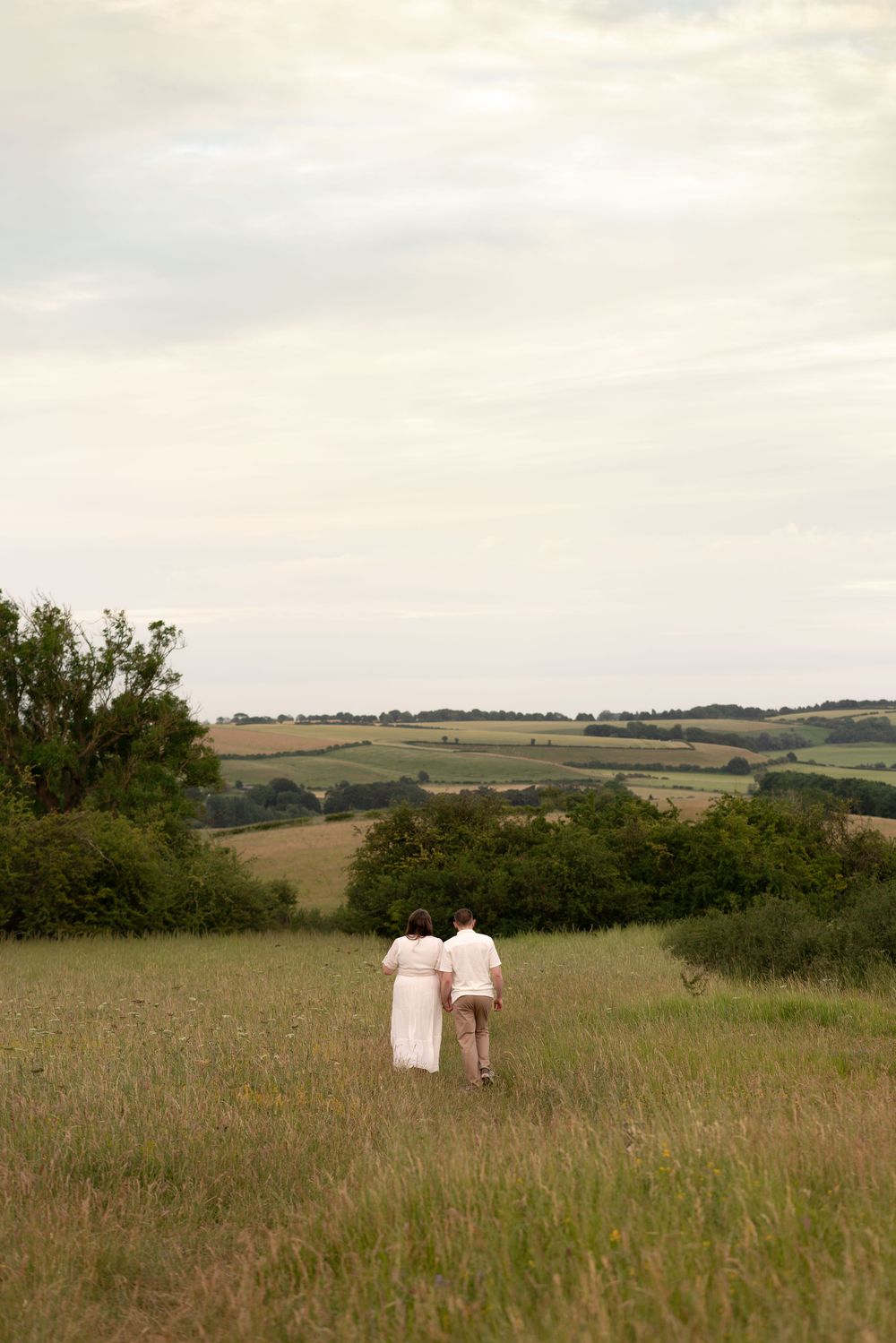 Couple in white walking through lush green meadow with rolling hills and trees under cloudy sky.