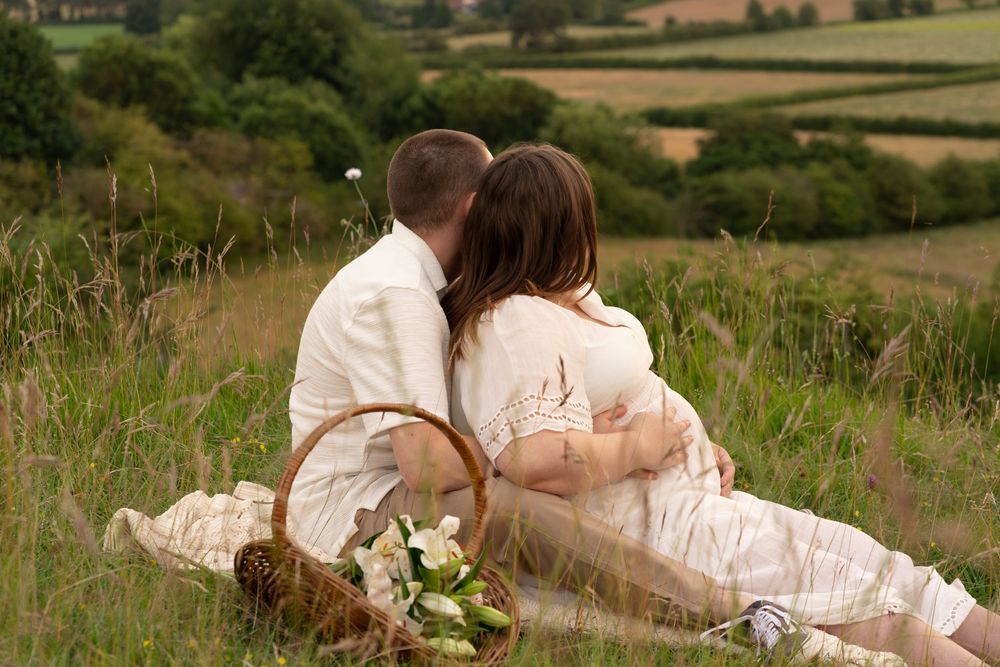 Romantic couple sharing a kiss during a countryside picnic on a grassy hill with a wicker basket and rolling fields.