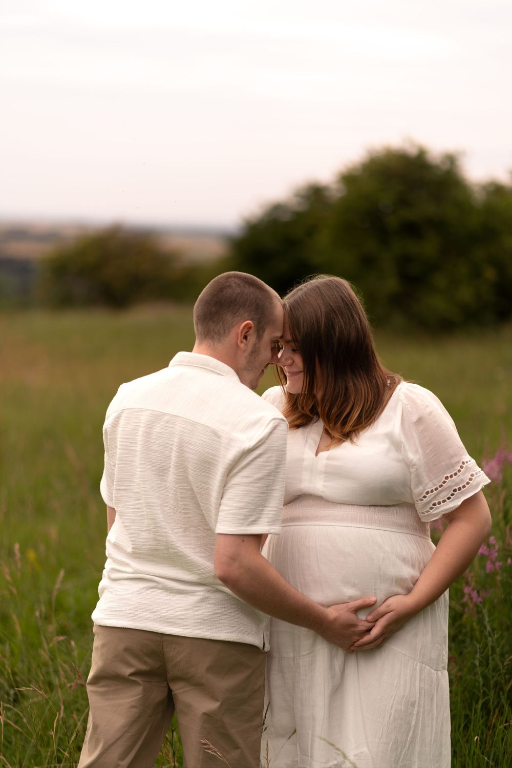 Couple sharing tender moment in green field, pregnant woman in white dress with partner touching her baby bump.