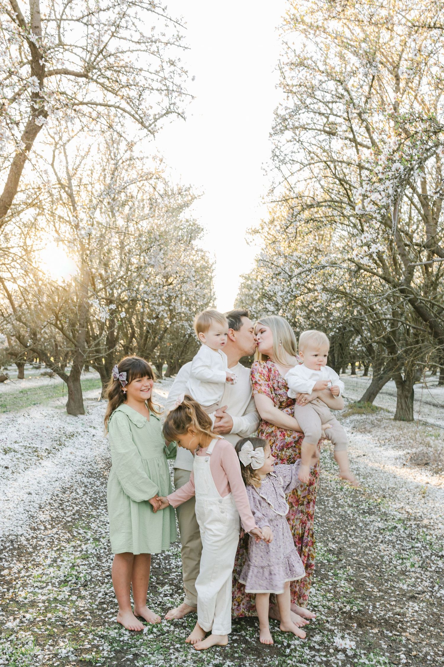 Family portrait in blooming orchard at golden hour, parents kissing while surrounded by four young children.