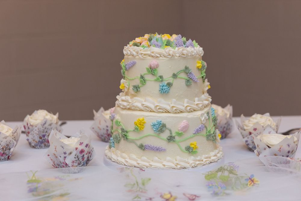 Three-tier white wedding cake with pastel floral decorations, surrounded by matching floral cupcakes on a white table.