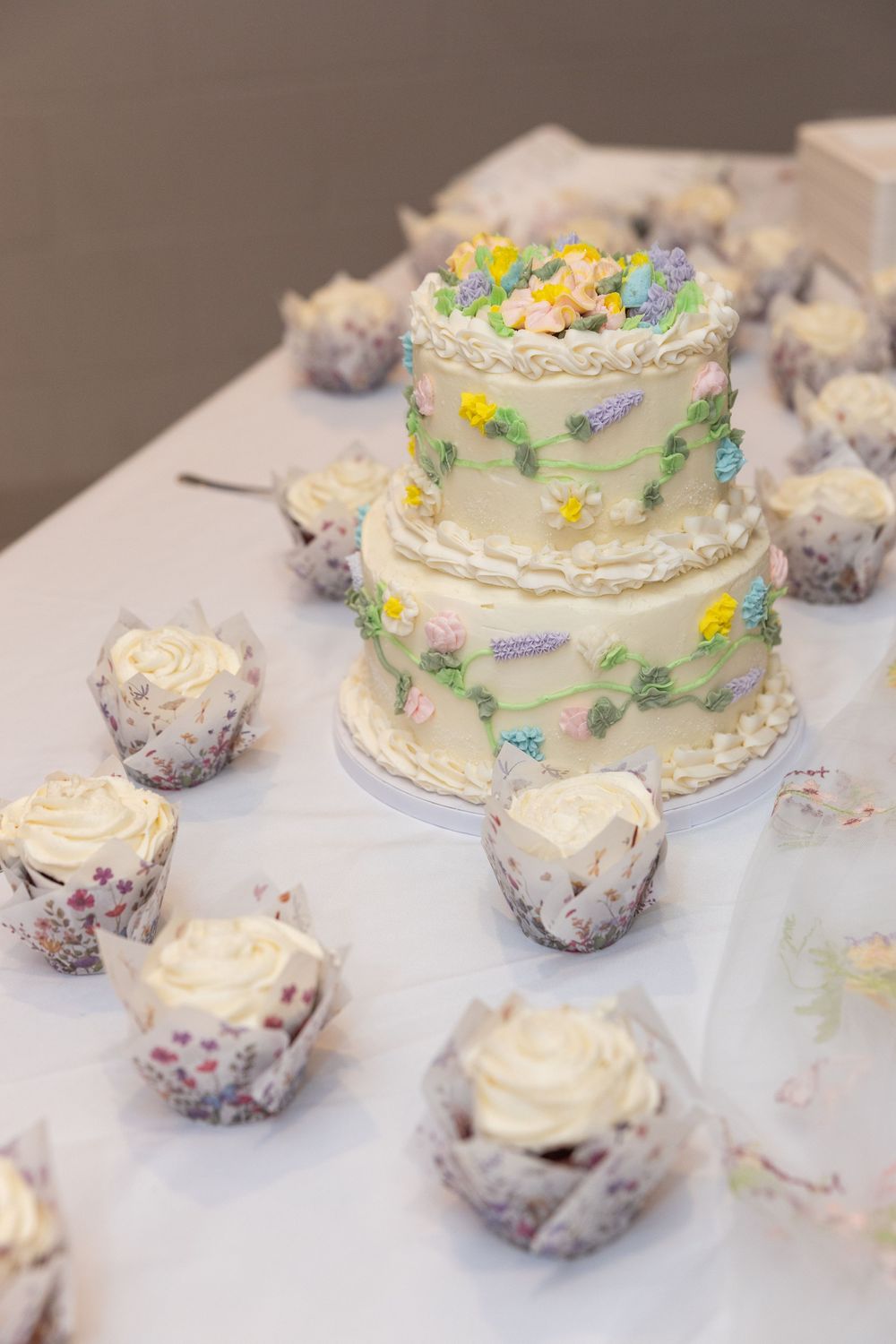 Elegant three-tier white wedding cake decorated with colorful flowers surrounded by cupcakes in floral wrappers on a white table.
