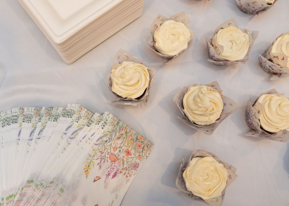 Vanilla frosted cupcakes in rustic paper liners displayed on a table beside floral napkins at a wedding reception.