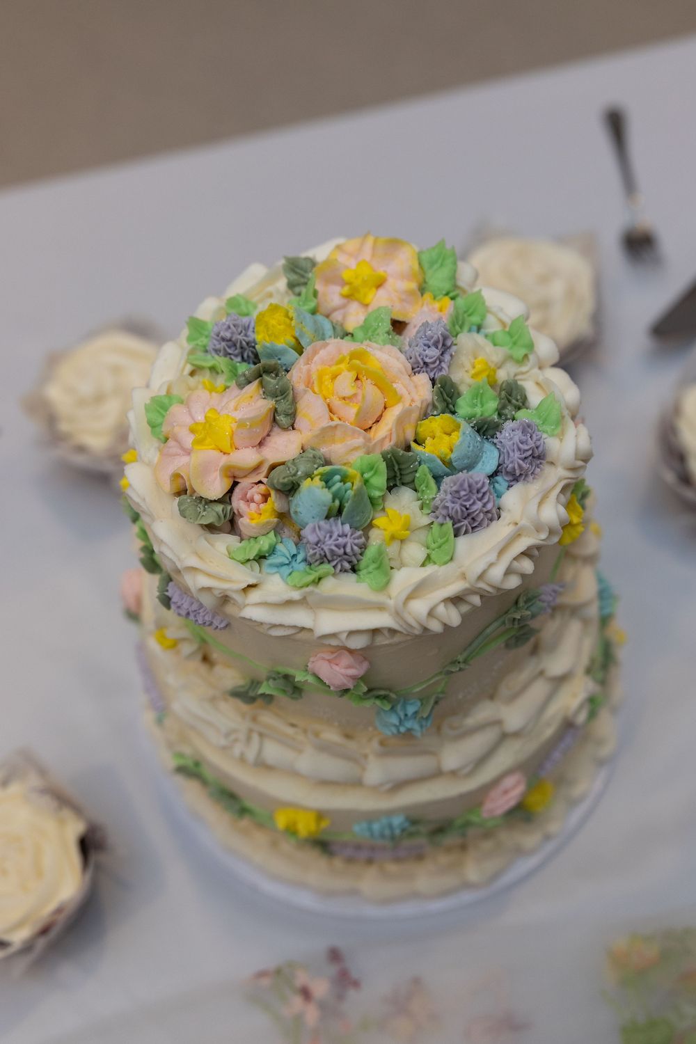 A two-tier naked wedding cake topped with pastel buttercream flowers including roses, lavender, and daffodils.