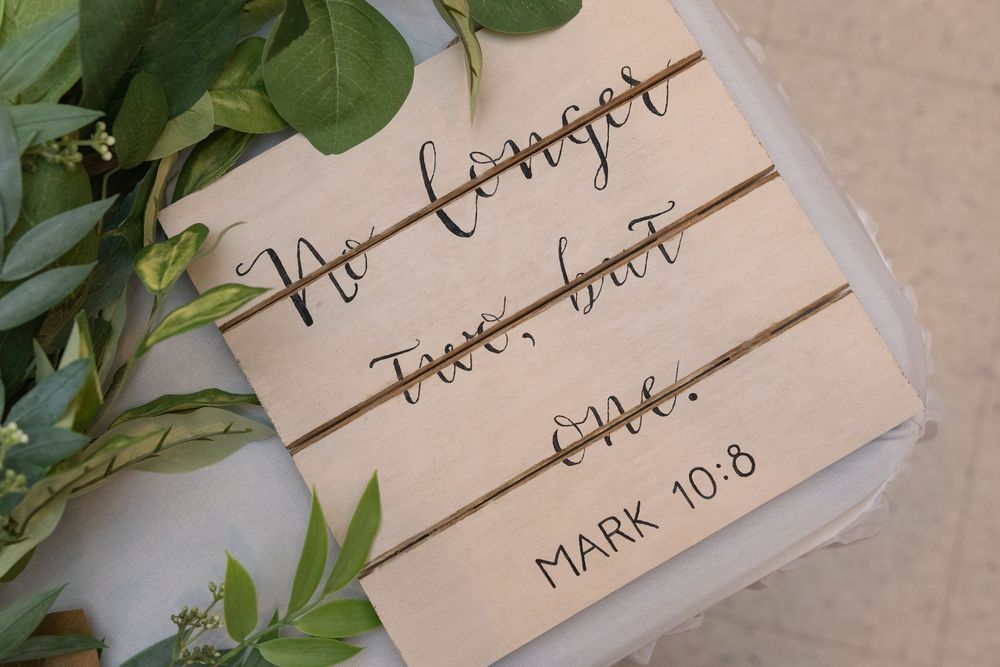 Wooden sign with cursive text reading No Longer Two But One with Bible verse Mark 10:8, surrounded by greenery.