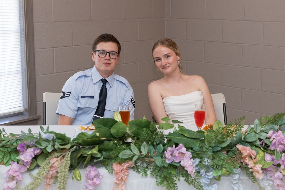 A young couple in formal attire sits at a decorated sweetheart table with floral garland and wine glasses.