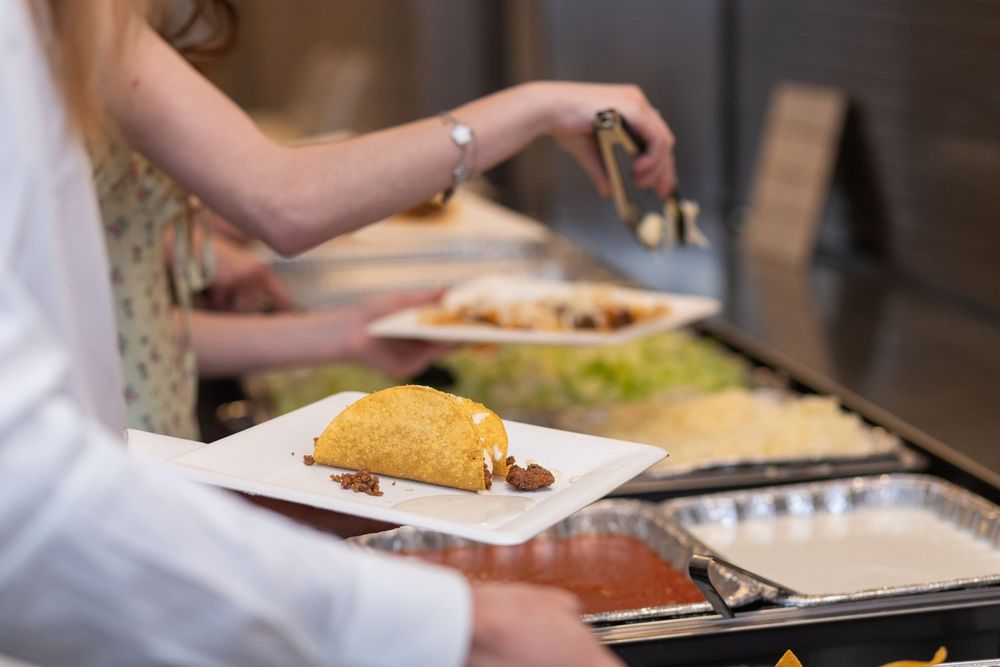 Guests serving themselves at a wedding buffet with taco and assorted dishes on white square plates.