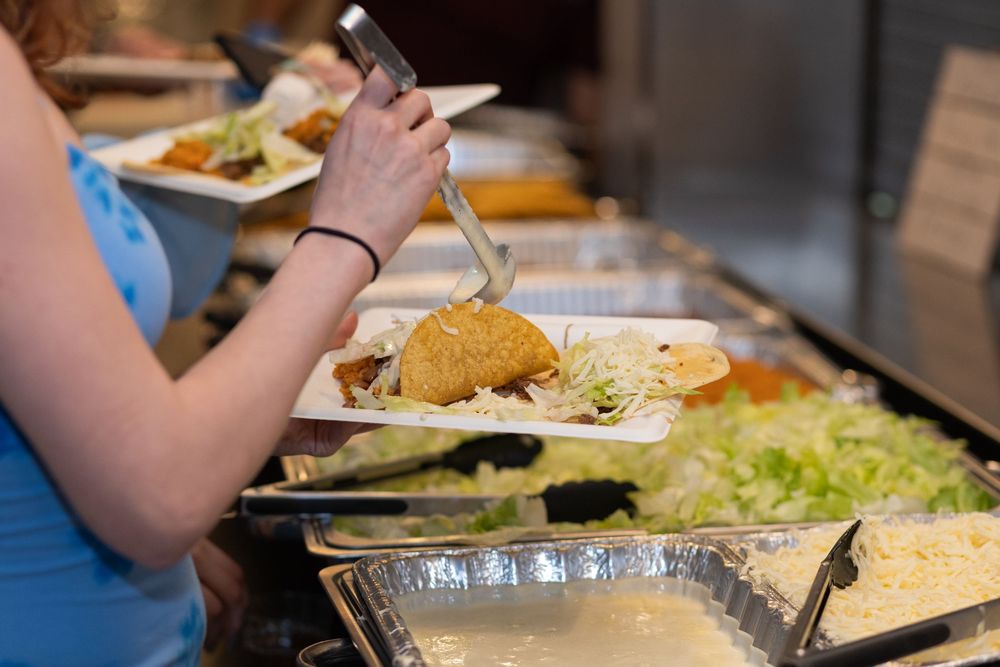 Woman serving herself food at a buffet line with various dishes including salad and fried items on white plates.