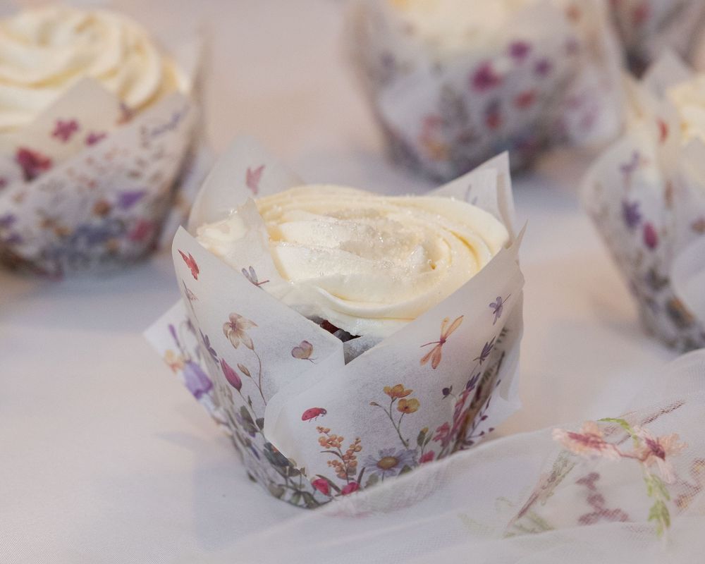 Close-up of a vanilla frosted cupcake in a decorative floral paper wrapper on a white surface.