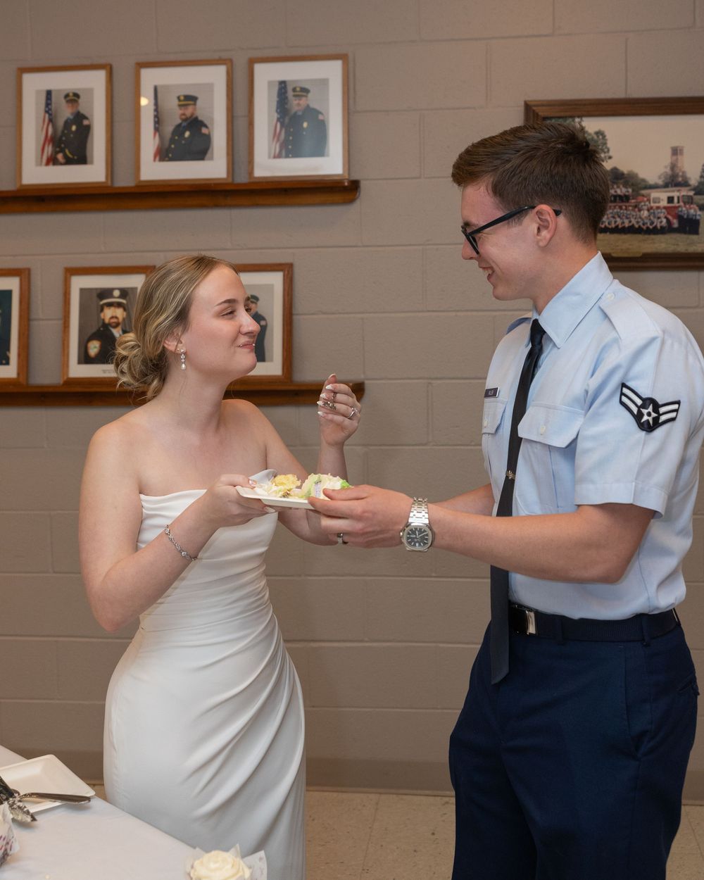 Military groom in Air Force uniform feeds wedding cake to bride in white dress at reception.