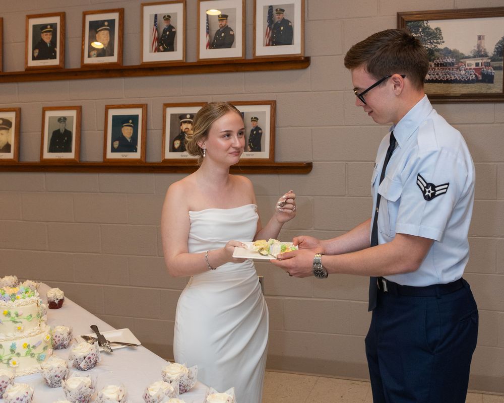 Military airman in uniform shares wedding cake with bride in white dress at reception with framed portraits on wall.