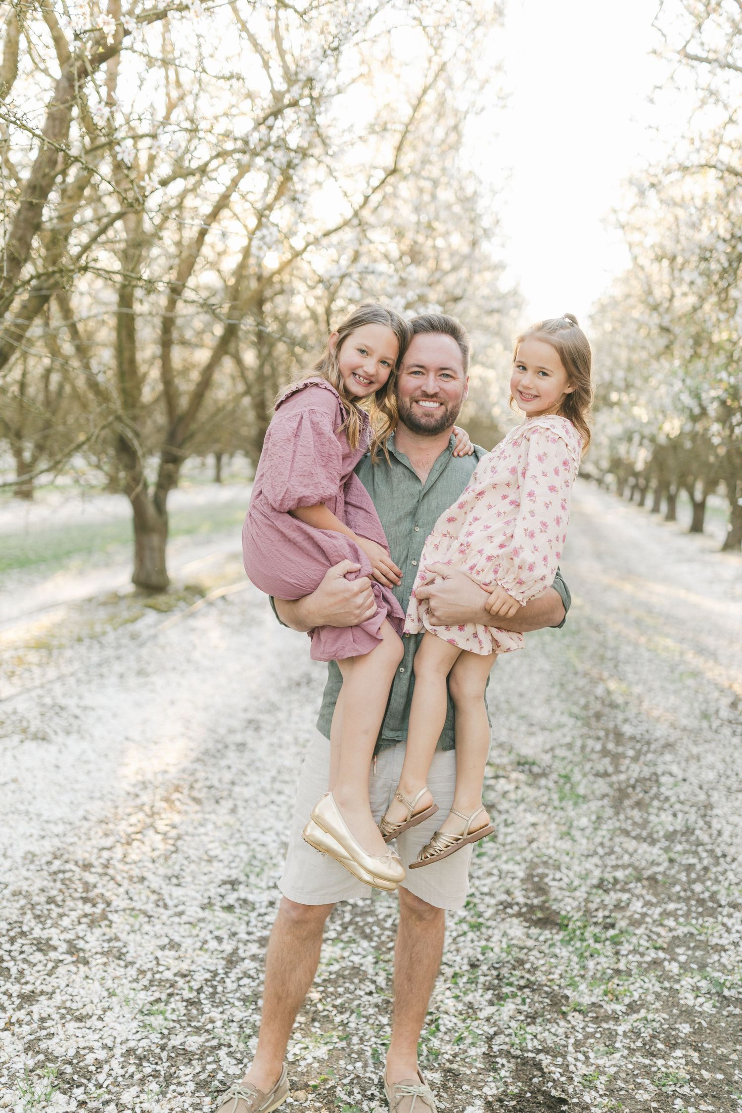 A smiling father holds two young daughters in pink dresses among blossoming trees on a petal-covered path.