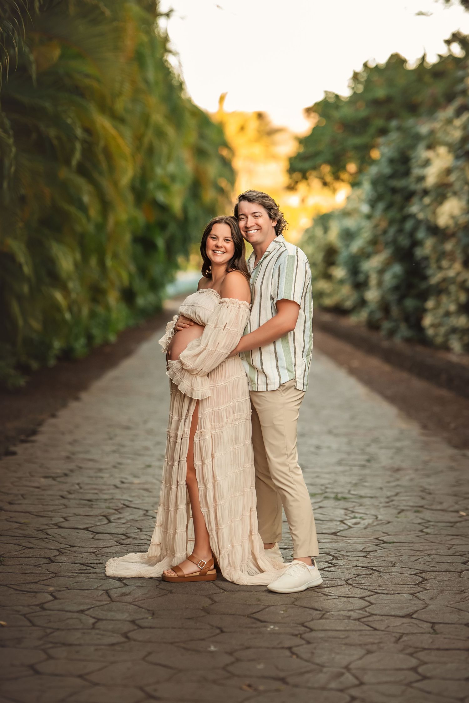 Smiling couple poses on a cobblestone path surrounded by lush greenery during a golden hour maternity photoshoot.