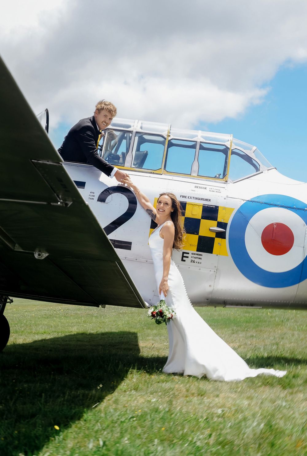 Bride in white gown holds groom's hand as he leans from vintage RAF aircraft cockpit on green airfield.
