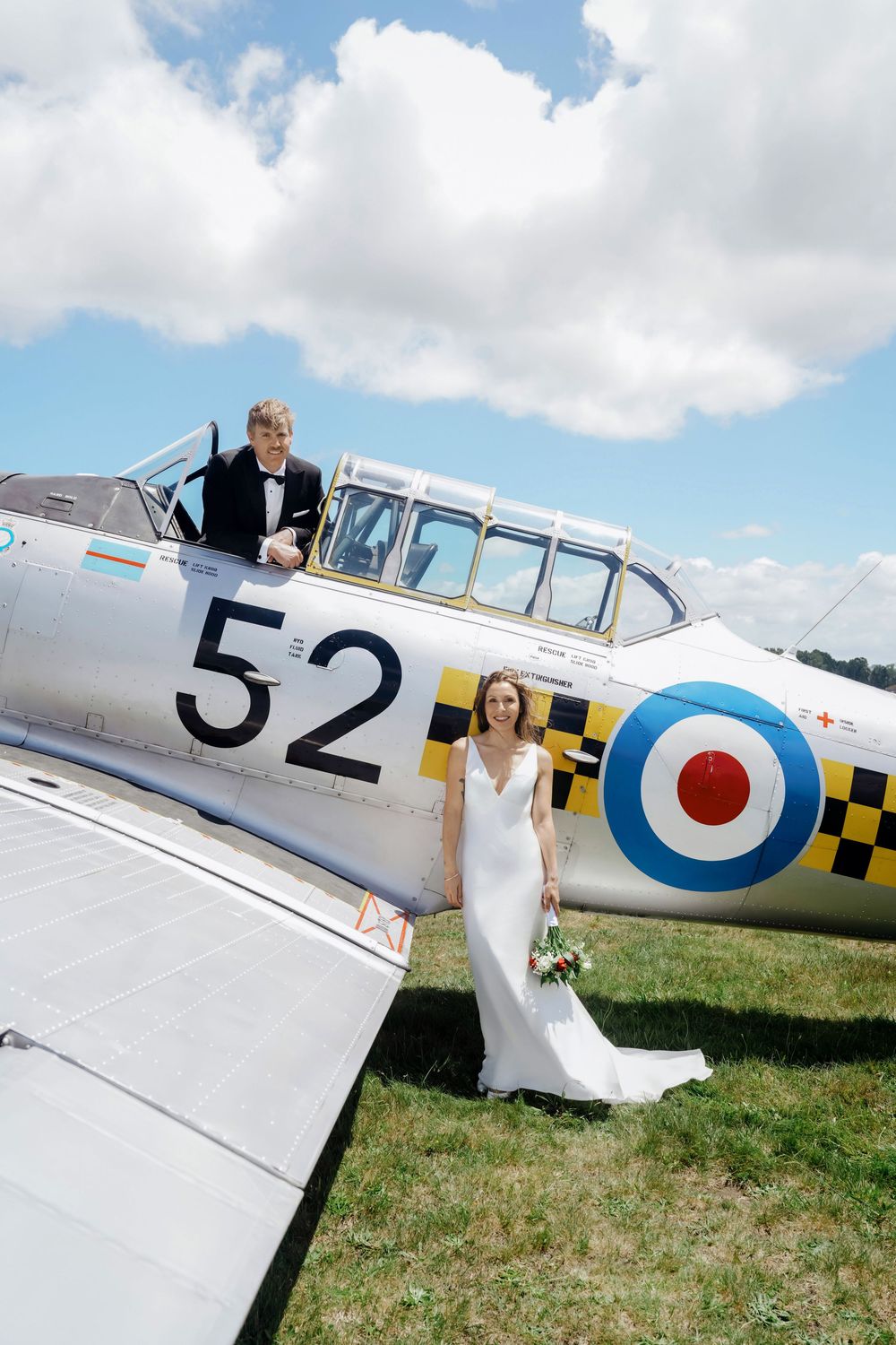 Bride in white gown stands by vintage WWII aircraft numbered 52 as groom in tuxedo sits in cockpit.