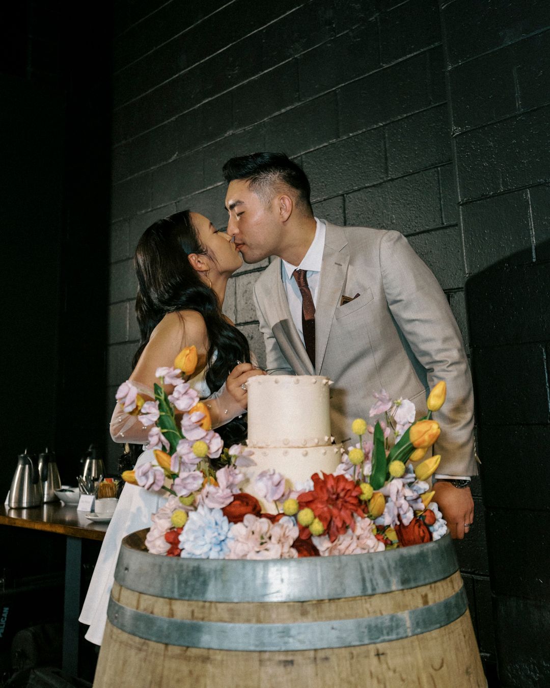 Couple kissing behind a floral-adorned wedding cake on a barrel, surrounded by colorful blooms at their wedding reception in Brisbane