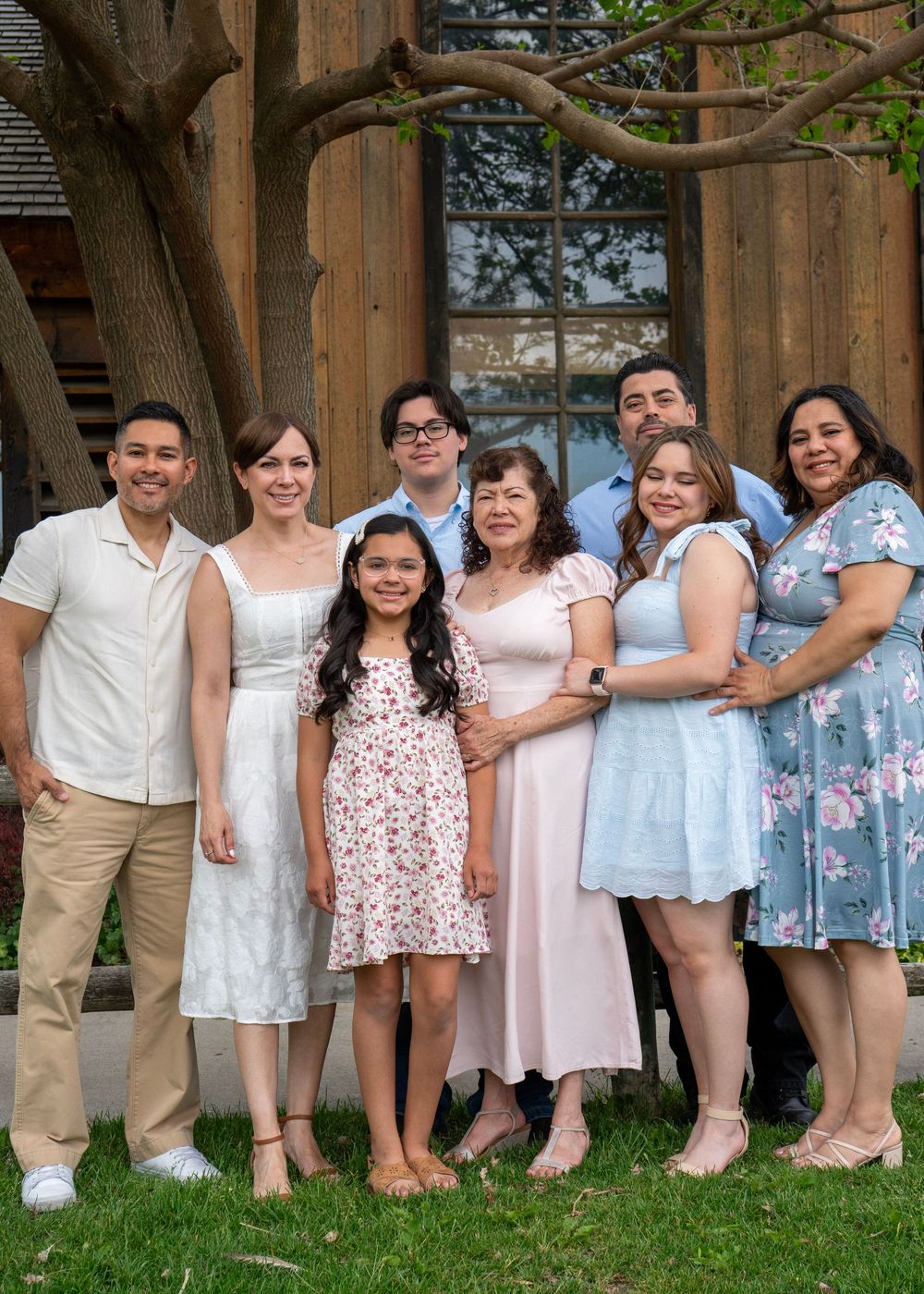 Large Hispanic family of seven smiling together outdoors in spring attire near a rustic wooden building.